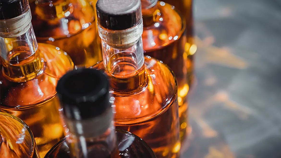unlabelled glass bottles filled with a golden-amber distilled spirit, such as whiskey, rum, or liquor, lined up during a production or bottling process