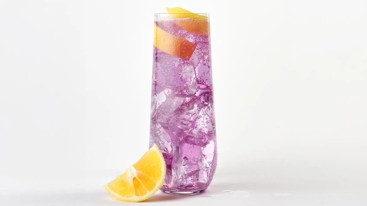 a refreshing purple cocktail in a faceted highball or champagne flute glass, garnished with citrus ribbons and served with ice and a lemon wedge on the side