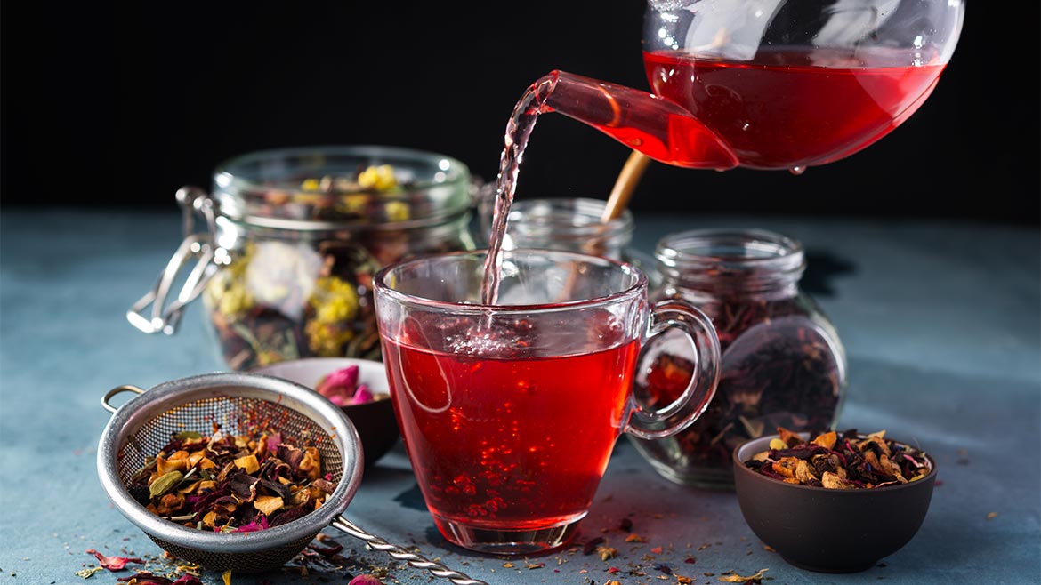 a vibrant, deep red herbal infusion—likely a hibiscus or fruit tea blend—being poured from a glass teapot into a matching glass cup