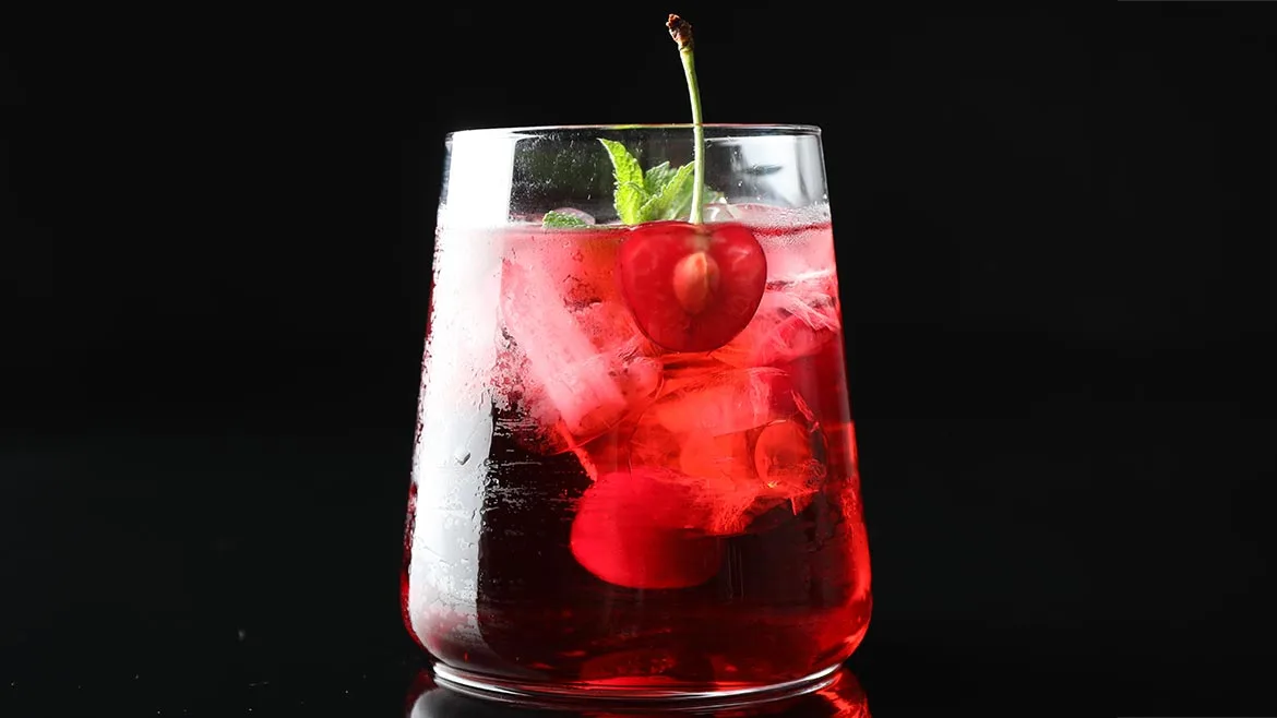 a refreshing red drink—such as a cherry soda water, cherry gin and tonic, or cherry mojito—filled with ice cubes, fresh cherries, and mint leaves against a dark background