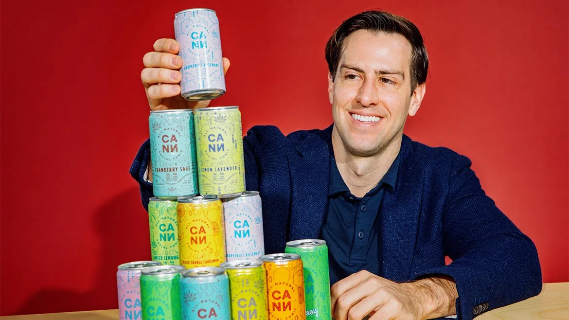 Jake Bullock, the co-founder and chief executive officer of the THC-infused social tonic brand Cann, posing alongside a stacked pyramid of various Cann beverage cans