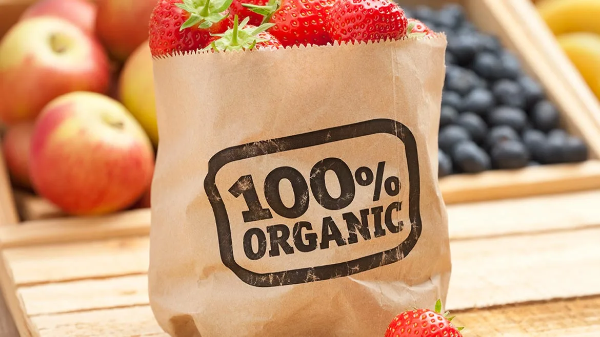 a brown paper bag filled with fresh strawberries, labeled "100% Organic," surrounded by other fruits like apples and blueberries.