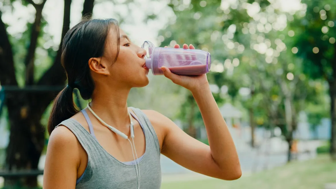 a person drinking a purple beverage from a shaker bottle outdoors near trees