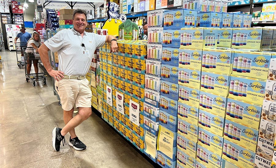 a man leaning against a large in-store display stack of Surfside Vodka Soda multi-packs inside a warehouse or grocery store